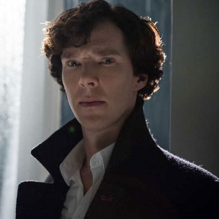 A celebrity in a dramatic pose, wearing a dark coat and standing inside a dimly lit room.