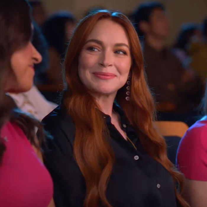 A woman with long red hair in a black shirt smiles, sitting between two others, in a casual setting.