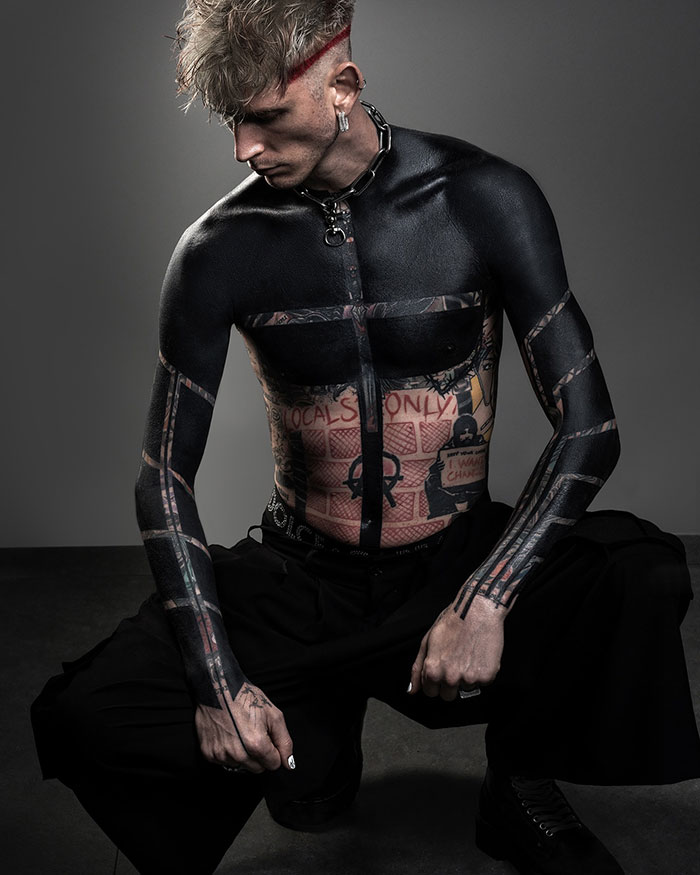 Machine Gun Kelly with blackout tattoo kneeling, showcasing intricate body art. Machine Gun Kelly with blackout tattoo kneeling, showcasing intricate body art.