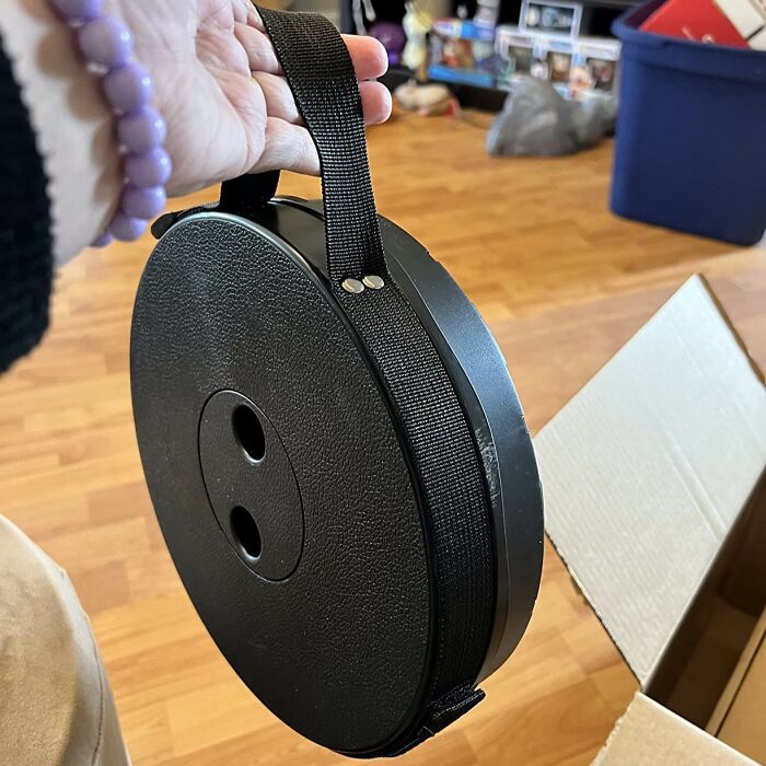 Hand holding a compact, circular black storage reel with a strap on a wooden floor, showcasing clever designs.