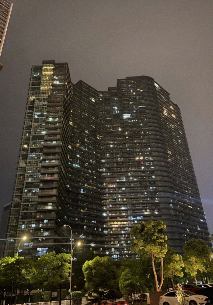 Night view of a large Chinese dystopian apartment building illuminated with many lights and surrounded by trees and cars.