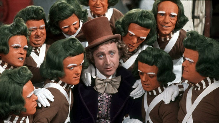 W***y Wonka surrounded by Oompa Loompas with green hair and orange faces in a classic movie scene about fun facts.