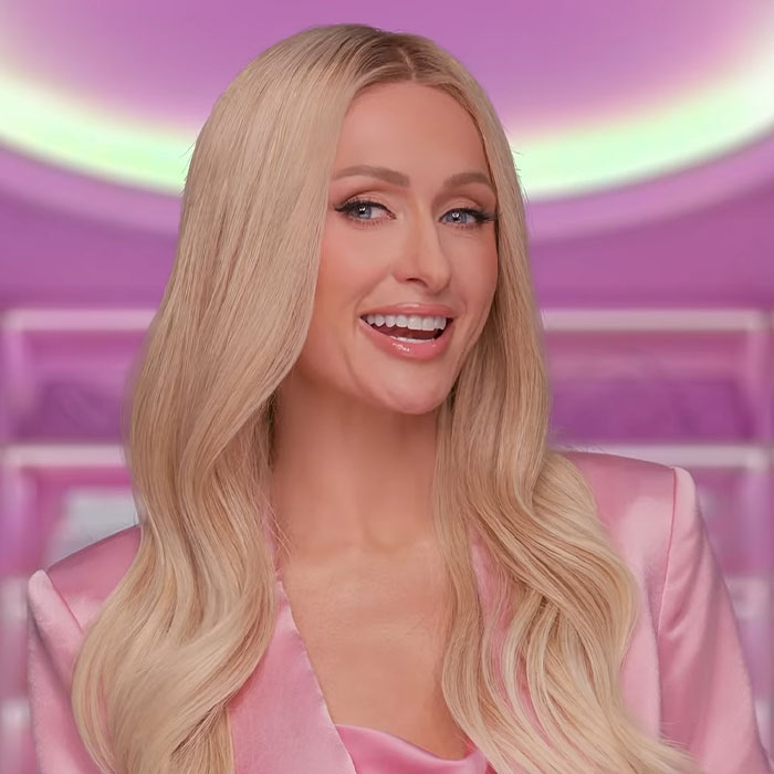 Blonde woman in a pink outfit, smiling in a vibrant setting related to celebrities and culture.