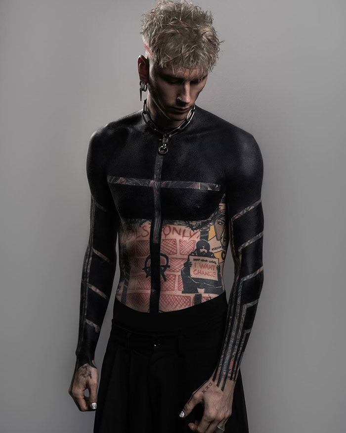 Machine Gun Kelly with a blackout tattoo on arms and chest, standing against a neutral background. Machine Gun Kelly with a blackout tattoo on arms and chest, standing against a neutral background.