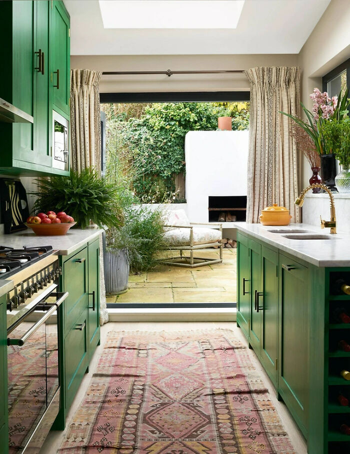 Galley kitchen with green cabinets, marble countertops, and large window opening to outdoor patio with greenery.