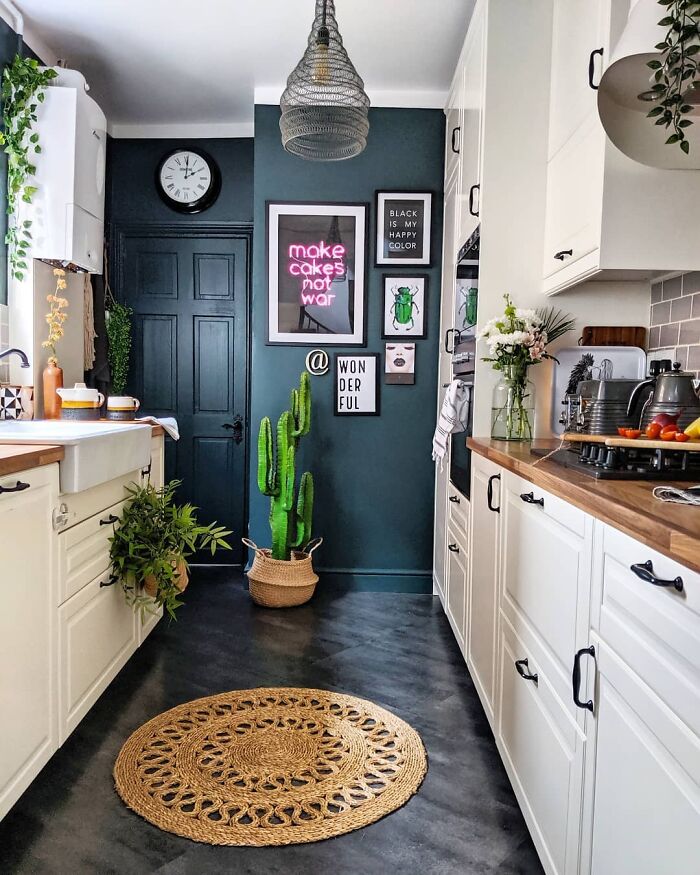 Cozy galley kitchen with white cabinets, wooden countertops, dark teal walls, plants, and decorative wall art.