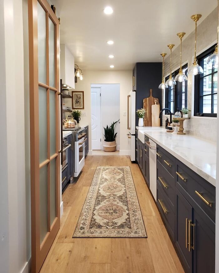 Galley kitchen with navy cabinets, brass hardware, white countertops, pendant lights, and a decorative runner on wood flooring.