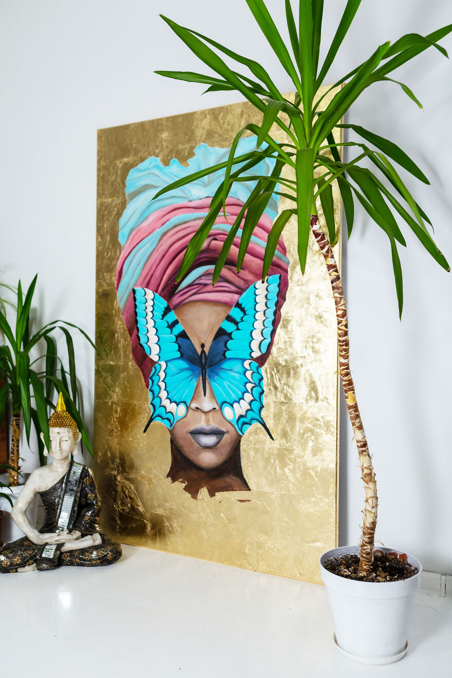 Painting of a woman with a colorful headwrap and blue butterfly covering her eyes, displayed near green plants indoors.