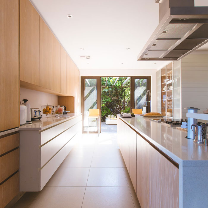 Bright modern galley kitchen with wooden cabinets and large windows opening to outdoor greenery and seating area.