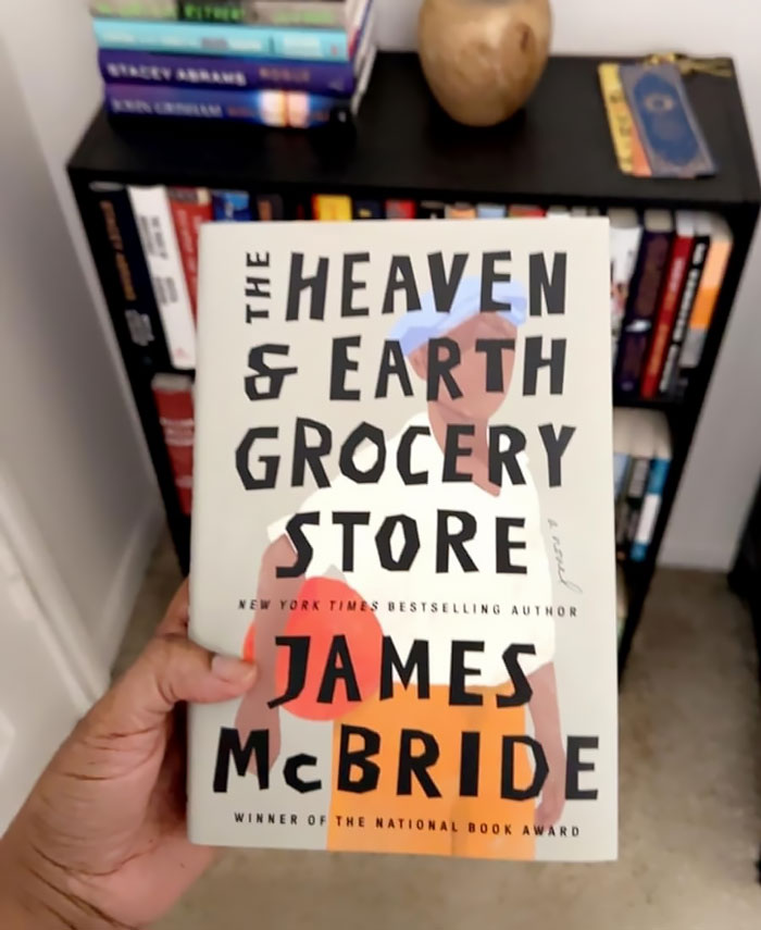 Hand holding "The Heaven & Earth Grocery Store" book, an ideal Galentine's Day gift for book lovers against a bookshelf backdrop.