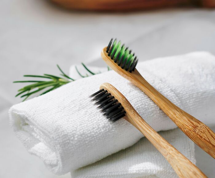 Two bamboo toothbrushes with black and green bristles resting on a white rolled towel and fresh greenery nearby.