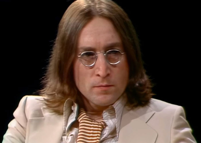 Man with long hair and round glasses wearing a beige suit and striped scarf, representing famous people declining major awards.