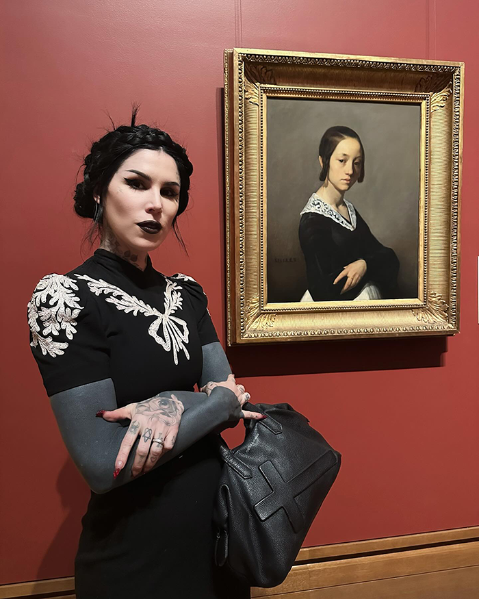 Megan Fox with tattoos standing by artwork, wearing a black dress with embroidered details, holding a black bag. Megan Fox with tattoos standing by artwork, wearing a black dress with embroidered details, holding a black bag.