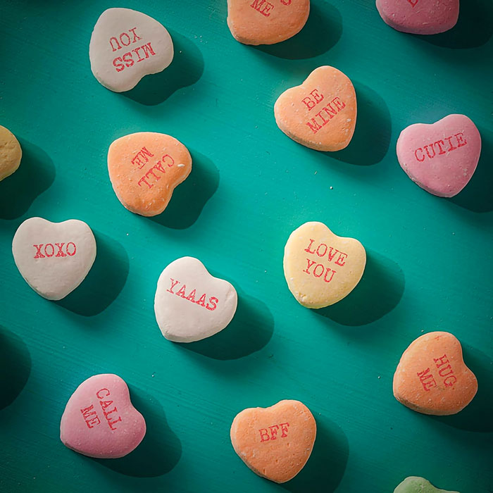 Colorful heart candies with Valentine's Day messages like "Be Mine" and "XOXO" on a teal background.