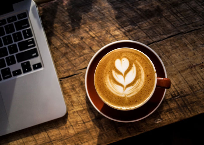 Cup of coffee with latte art on a wooden table next to a laptop, illustrating relatable embarrassing late-life confessions.