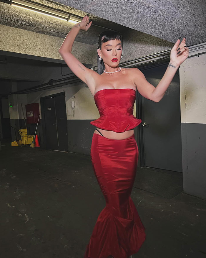Katy Perry in a red gown showing a black thong and arm tattoo, posing with arms raised.