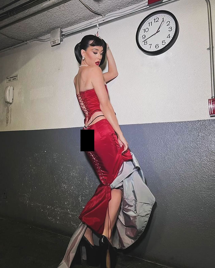 Katy Perry in a red dress poses in a hallway under a clock, showcasing a butterfly tattoo.