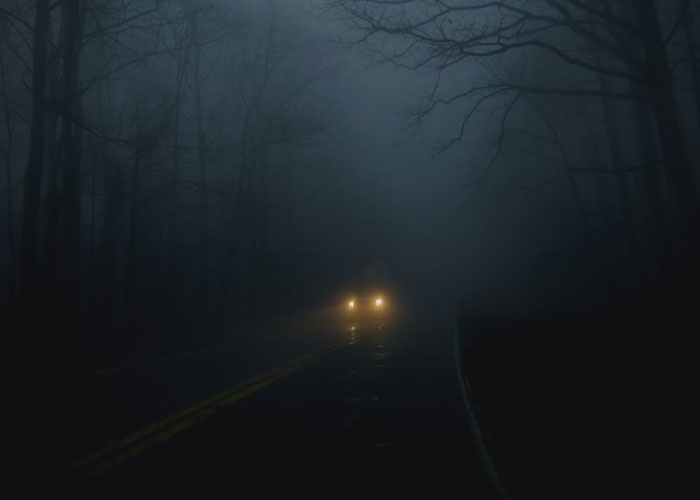 Car headlights shining through dense fog on a dark forest road, capturing an eerie and mysterious atmosphere.