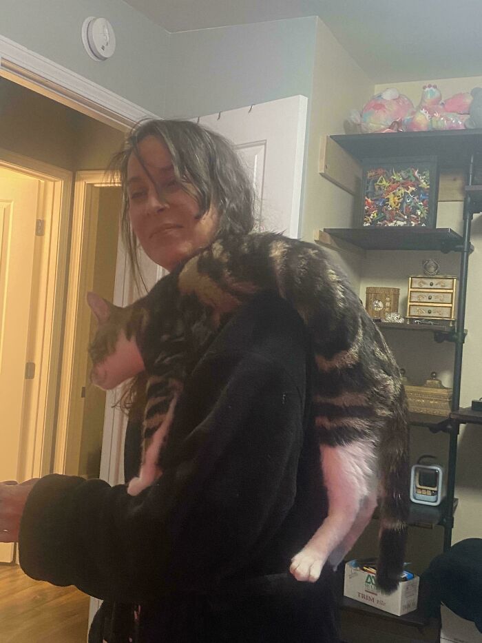 Woman in a black robe holding a malfunctioning cat draped awkwardly over her shoulder indoors at home.