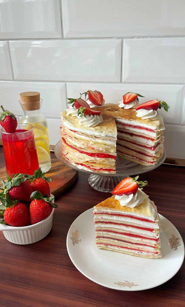 Layered crepe cake with strawberries and cream, fresh strawberries in a bowl, and red strawberry drink on kitchen counter.