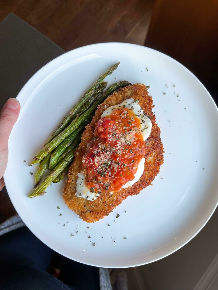 Hand holding a white plate with a perfectly breaded and sauced food serving accompanied by grilled asparagus vegetables.