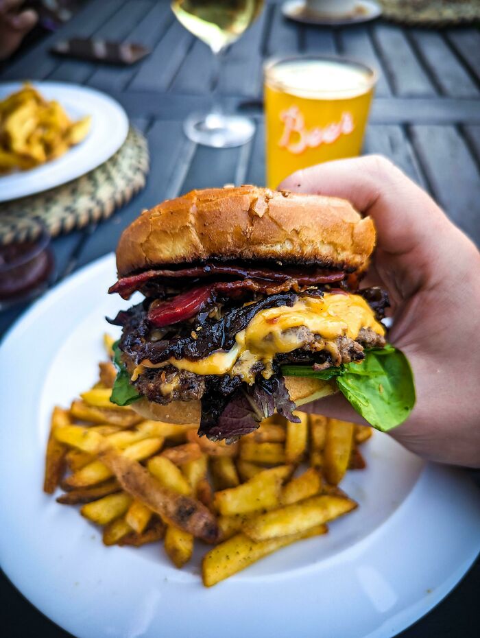 Close-up of a perfectly crafted cheeseburger with bacon and lettuce, held above a plate of golden French fries.
