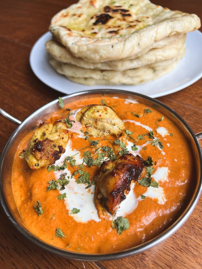 Butter chicken garnished with cream and herbs served with stacked naan bread on a white plate, food perfection achieved.
