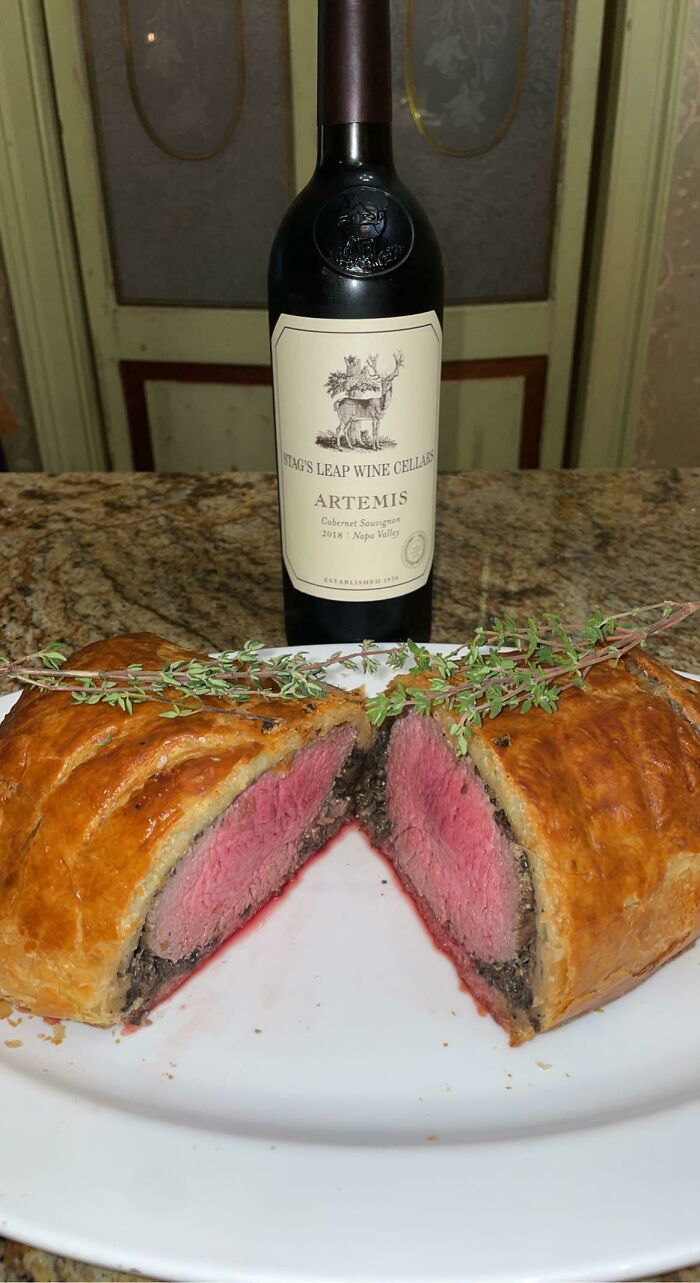 Perfectly cooked beef Wellington sliced open on a plate with fresh herbs and a bottle of red wine in the background.
