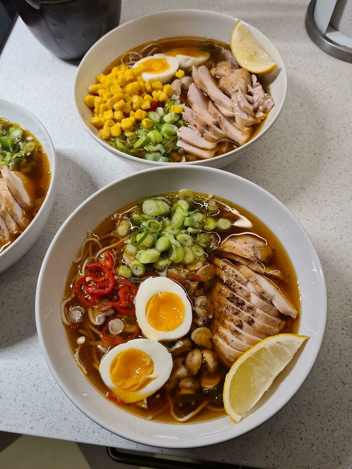 Two bowls of perfectly prepared ramen with sliced chicken, soft-boiled eggs, corn, green onions, chili, and lemon wedge on a countertop.