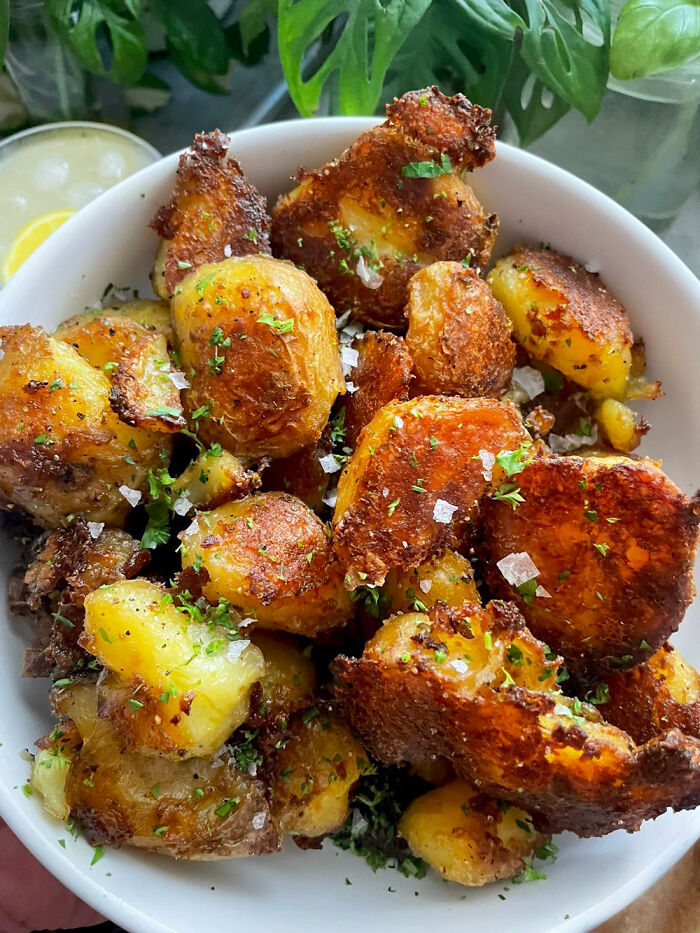 Crispy smashed potatoes seasoned with herbs and sea salt, showcasing food perfection in a white bowl with greenery in the background.