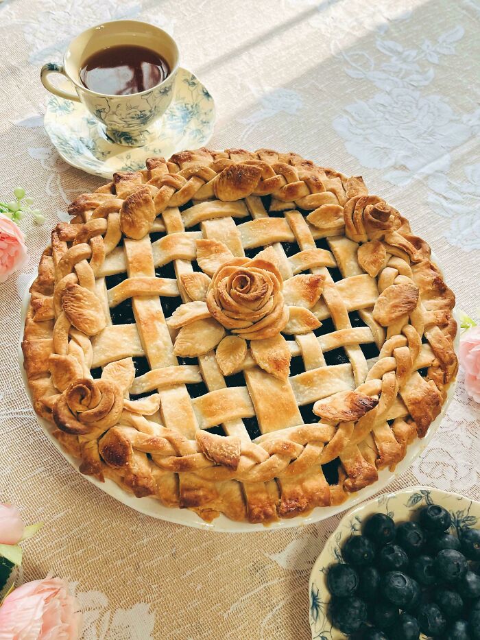 Perfectly crafted lattice blueberry pie with decorative crust, served with tea and fresh blueberries on a floral tablecloth.