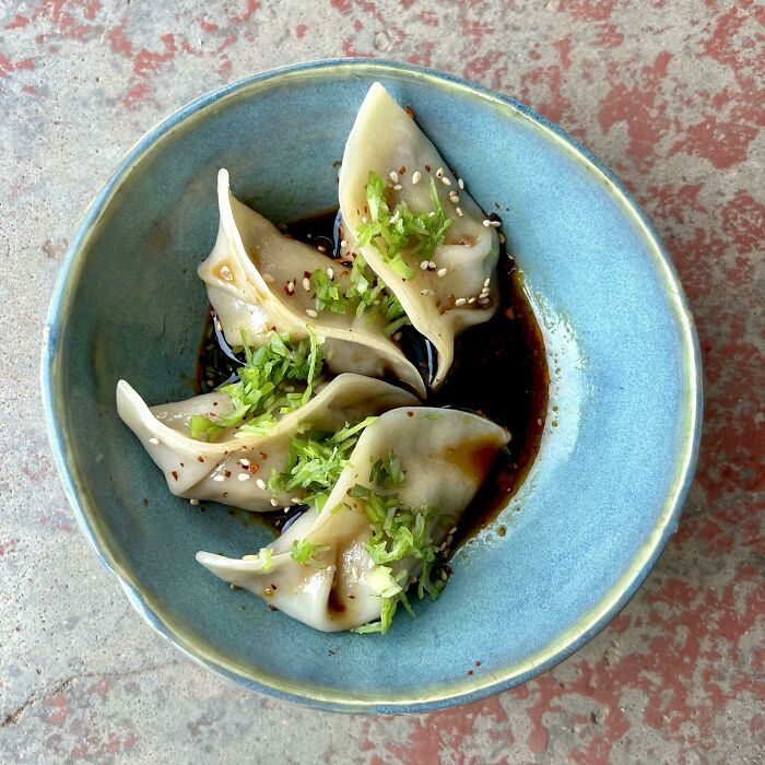 Four perfectly shaped dumplings garnished with herbs and sesame seeds served in soy sauce in a blue bowl.