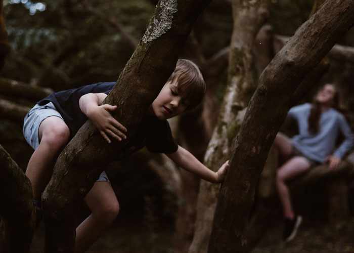 Young boy climbing a tree in a forest with a blurred older person in the background, reflecting childfree people over 40 themes.