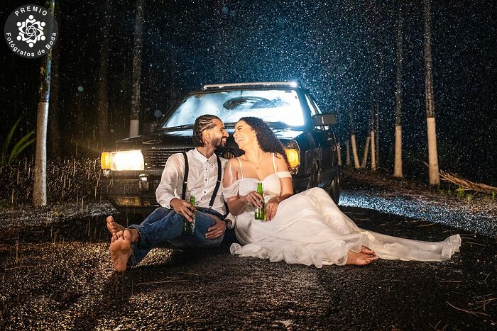 Couple laughing barefoot in the rain, sitting by a car, holding bottles, showcasing an incredible wedding moment.
