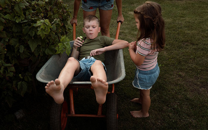 Child sitting in a wheelbarrow while a woman and a girl stand nearby, representing summer babysitting situation.