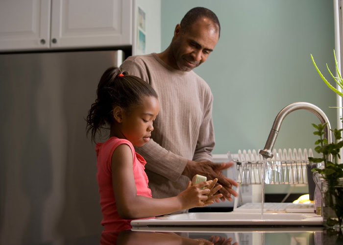 Man helps young girl wash hands in kitchen, reflecting a moment of care and family connection.