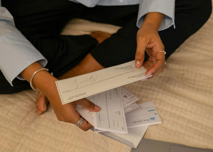 Woman holding multiple checks while sitting cross-legged on a bed, considering no-contact after lawsuit payout demand
