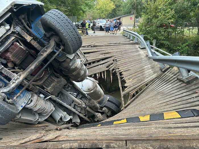 Truck overturned on a collapsed wooden bridge while bystanders observe the clueless accident scene outdoors.