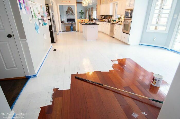 Partially painted wooden floor in kitchen showing a DIY disaster with uneven white paint coverage and tools left on floor