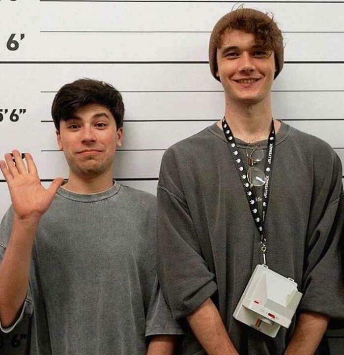 Two people in oversized gray shirts stand in front of a height chart, one waving. Comedy and youth captured in style.