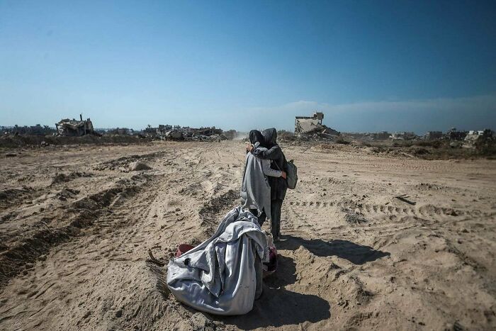 Two people hug on barren land, suggesting a boring dystopia with desolate surroundings and distant ruined buildings.