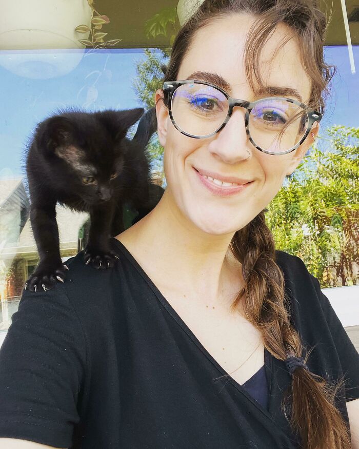 A woman smiling with a black cat on her shoulder, showing its sharp claws in an adorable pose.