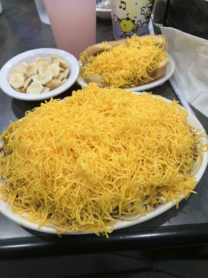 Extra cheese piled on a Cincinnati-style chili plate, accompanied by a hot dog with additional cheese and a bowl of crackers.