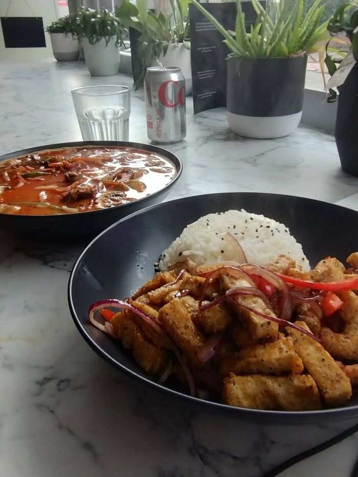 Tofu stir-fry with rice and extra sauce, served with a Diet Coke, in a cafe setting with plants on the table.