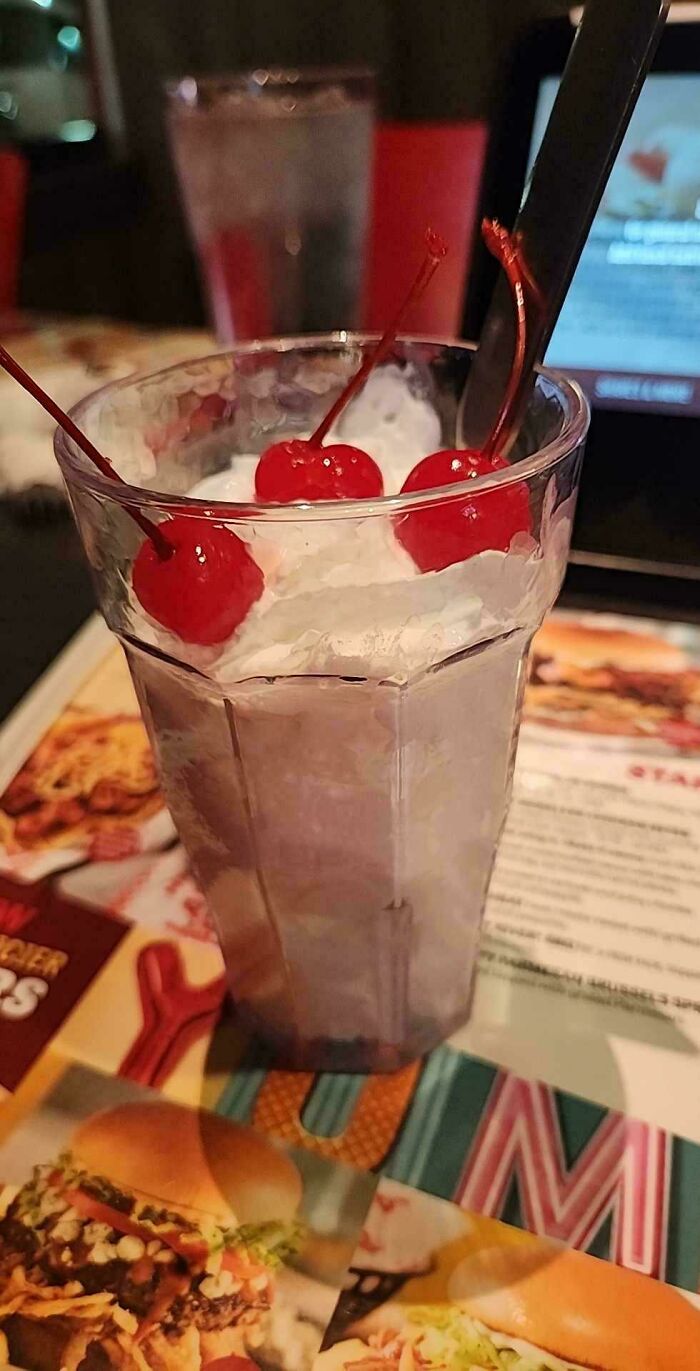 Milkshake topped with extra cherries, served in a tall glass, on a table with a menu in the background.