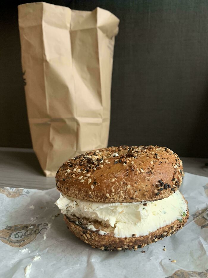 Bagel loaded with extra cream cheese, complying with a food service request, placed on a paper wrapping next to a brown bag.