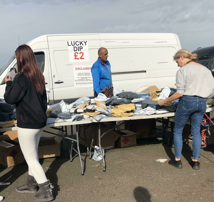 People browsing a table of packages at a UK market, with a van labeled “Lucky Dip £2” in the background.