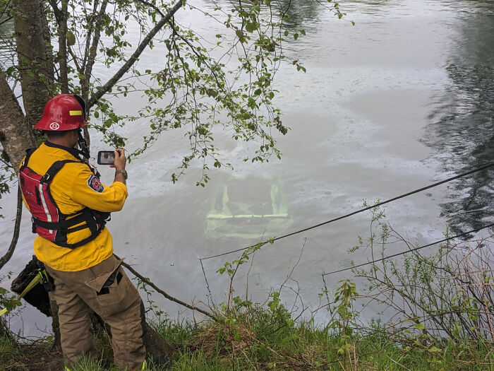 Incident scene with a rescue worker photographing a submerged vehicle in a river, suggesting a costly mishap.