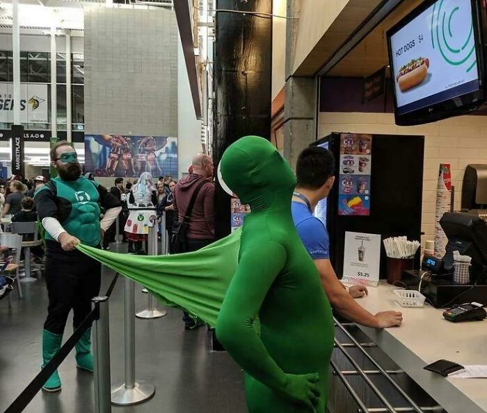 Two people in superhero costumes at a food counter, with one tugging on the other's green outfit playfully.