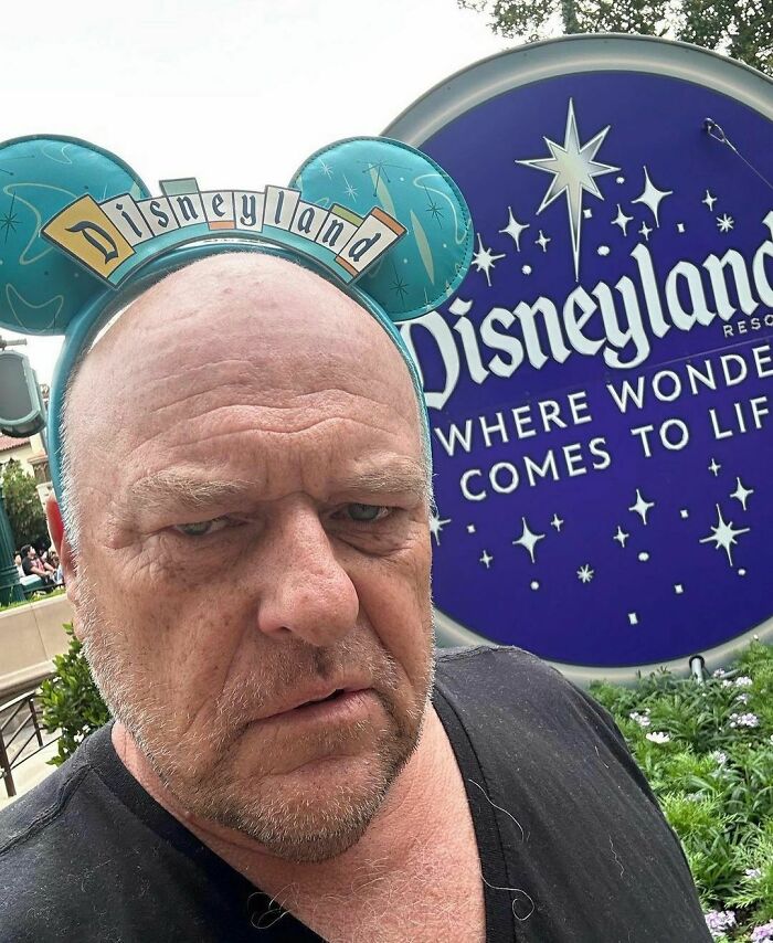 Man wearing Disneyland Mickey ears, looking perplexed in front of a Disneyland sign; something unusual catches his attention.